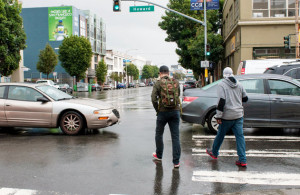 Pedestrians crossing the street while walking around a car that is blocking the box.